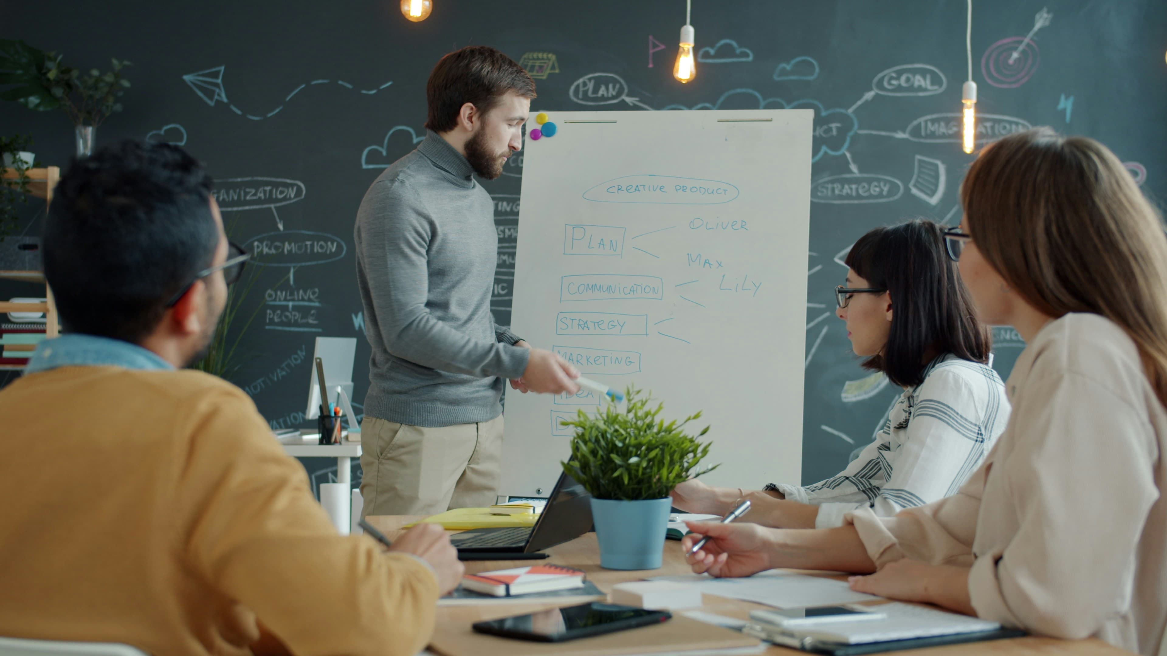 Team gathered around a whiteboard in a modern office, one person presenting and writing while colleagues discuss strategy