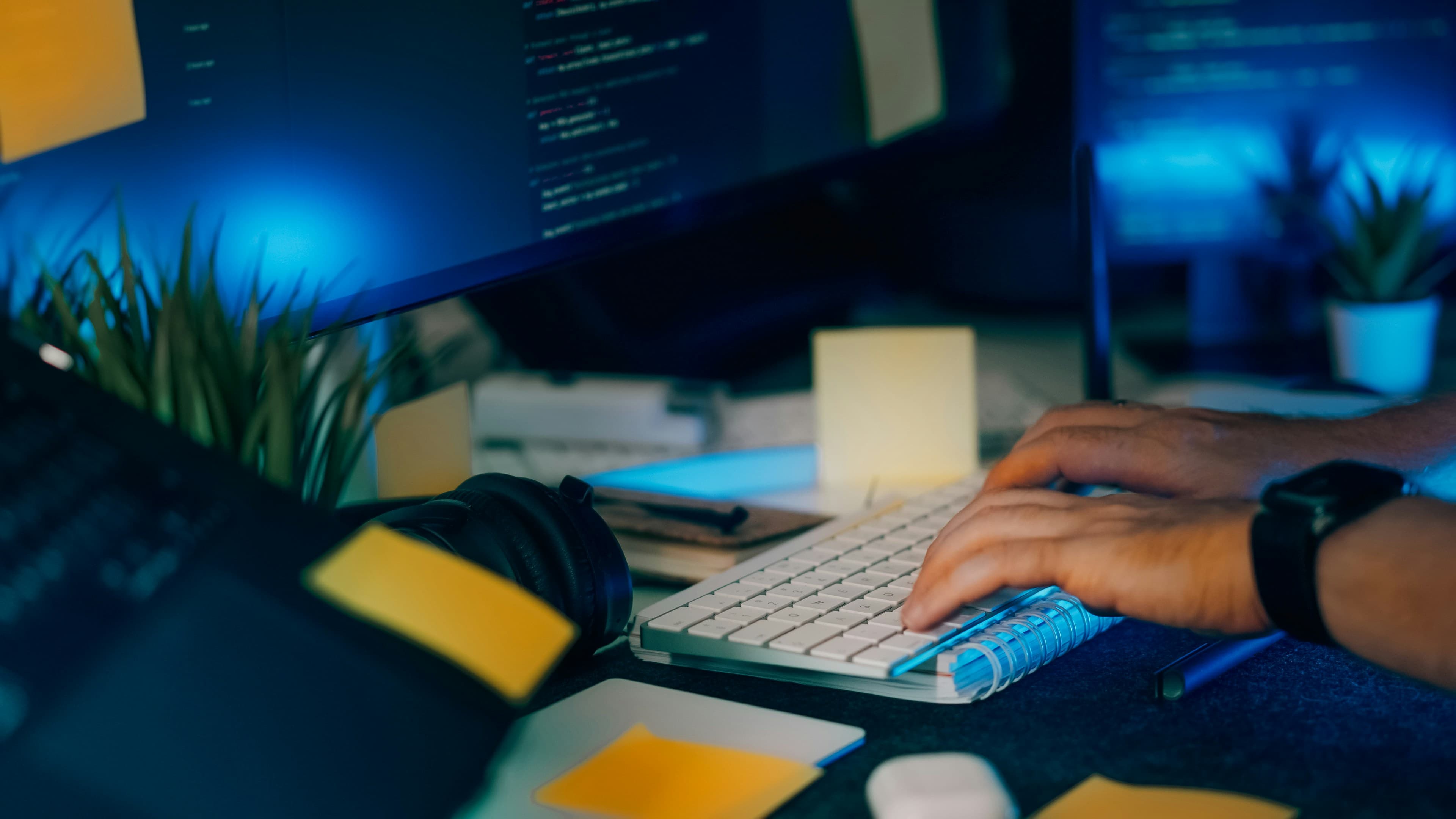 Close-up of hands typing on a keyboard with computer screens showing code in the background