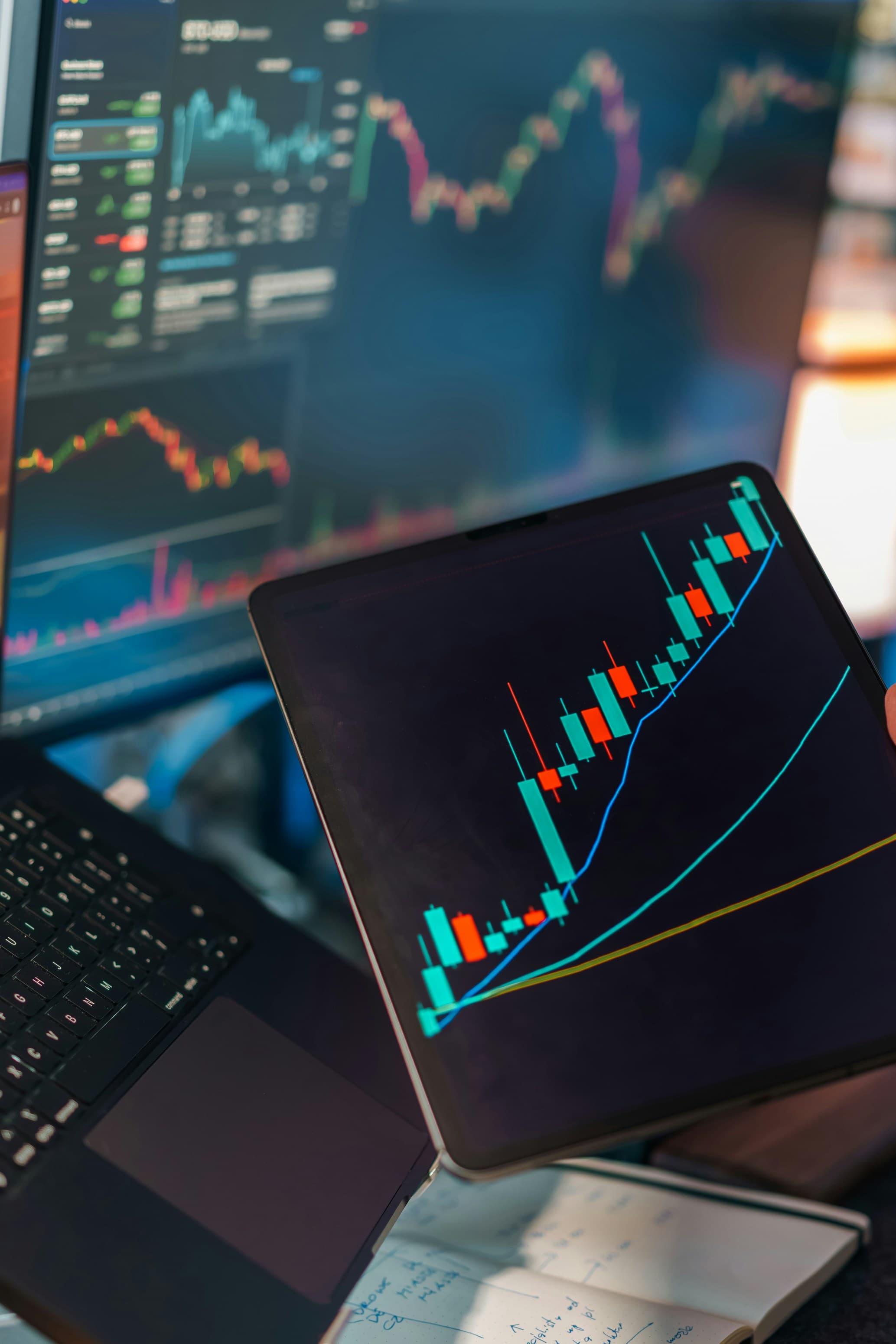 Financial analytics workspace with laptop and tablet displaying candlestick charts and technical data visualizations